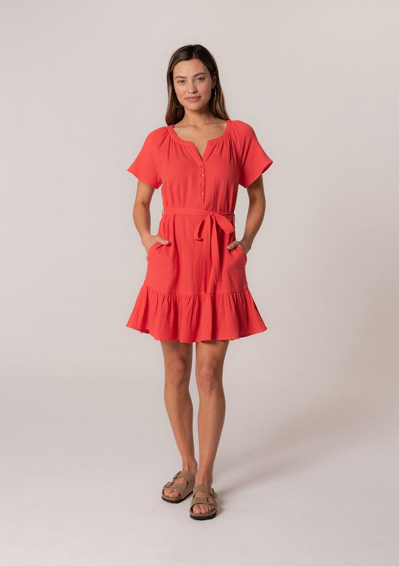 [Color: Flame] A full body front facing image of a brunette model wearing a spring solid red mini dress designed in cotton gauze. With short sleeves, a button front top, a v neckline, a tiered mini skirt, and a tie waist belt. 