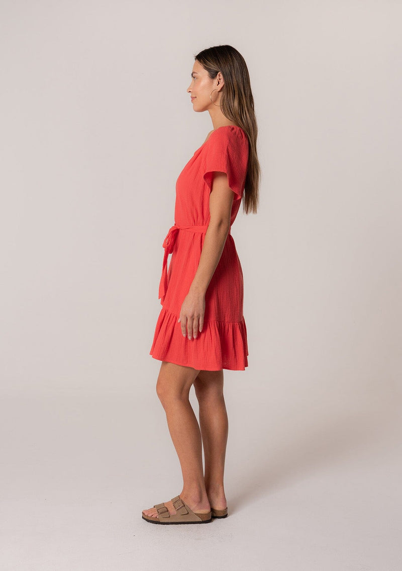 [Color: Flame] A side facing image of a brunette model wearing a spring solid red mini dress designed in cotton gauze. With short sleeves, a button front top, a v neckline, a tiered mini skirt, and a tie waist belt. 