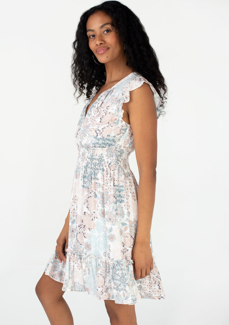 [Color: Dusty Blue/Teal] A side facing image of a brunette model wearing a bohemian spring mini dress in a blue floral print. With short flutter sleeves, a smocked elastic waist, a flowy ruffle trimmed tiered skirt, and a v neckline. 