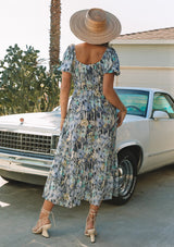 [Color: Dusty Lilac/Dusty Teal] A back facing image of a blonde model standing outside in front of a vintage white car wearing a spring bohemian maxi dress in a blue floral print. With short puff sleeves, a square neckline, an elastic waist, and a tiered skirt.