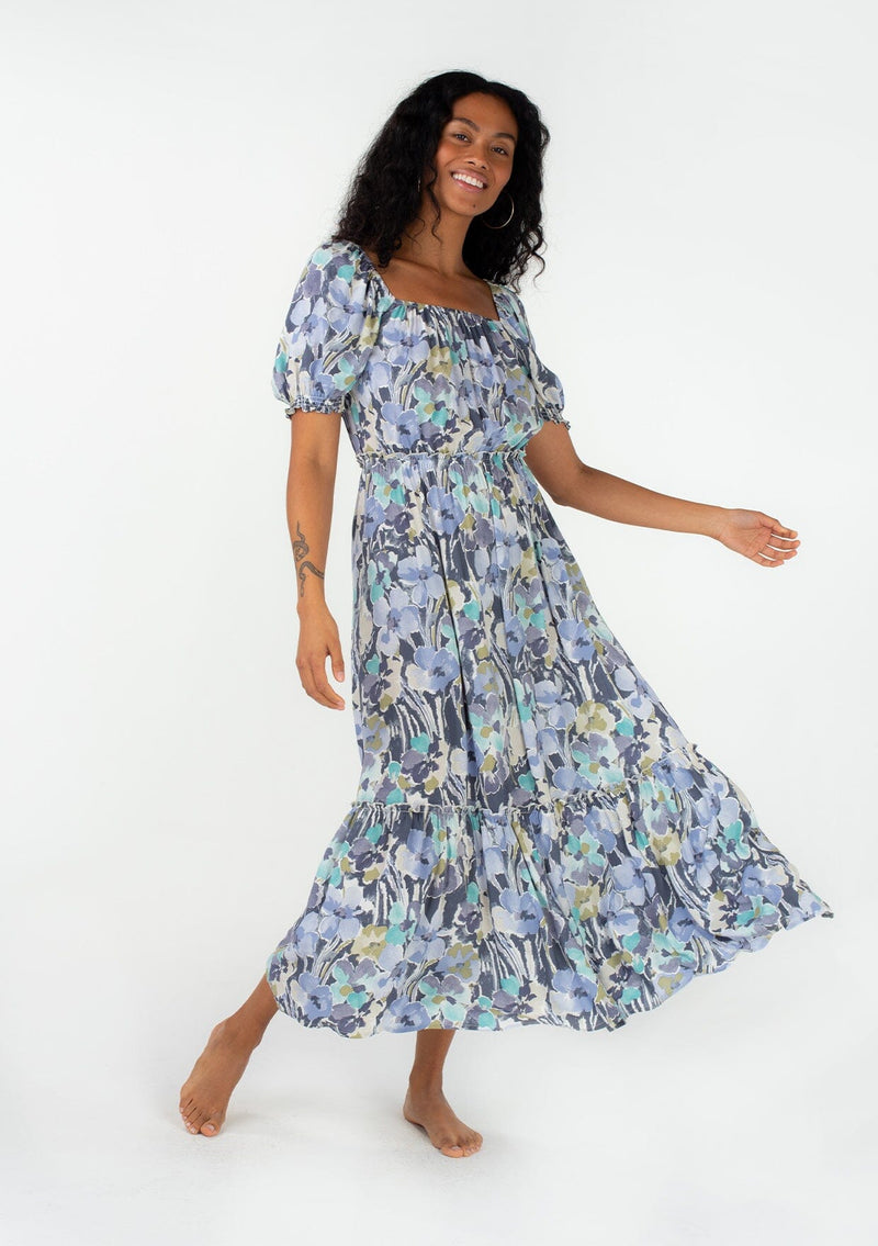 [Color: Dusty Lilac/Dusty Teal] A full body front facing image of a brunette model wearing a spring bohemian maxi dress in a blue floral print. With short puff sleeves, a square neckline, an elastic waist, and a tiered skirt. 