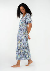 [Color: Dusty Lilac/Dusty Teal] A side facing image of a brunette model wearing a spring bohemian maxi dress in a blue floral print. With short puff sleeves, a square neckline, an elastic waist, and a tiered skirt. 