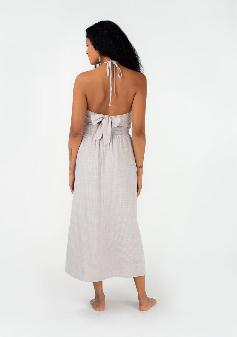[Color: Dove] A back facing image of a brunette model wearing a spring halter maxi dress in a light grey cotton seersucker check. With a drawstring halter neckline that ties at the back, side pockets, a half smocked elastic waistline, and an adjustable back tie detail. 