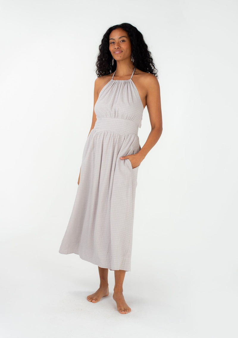 [Color: Dove] A front facing image of a brunette model wearing a spring halter maxi dress in a light grey cotton seersucker check. With a drawstring halter neckline that ties at the back, side pockets, a half smocked elastic waistline, and an adjustable back tie detail. 
