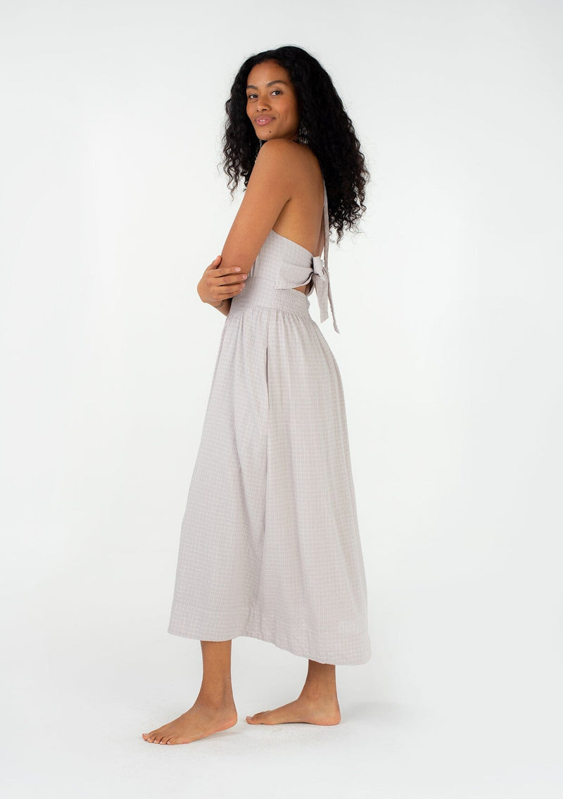 [Color: Dove] A side facing image of a brunette model wearing a spring halter maxi dress in a light grey cotton seersucker check. With a drawstring halter neckline that ties at the back, side pockets, a half smocked elastic waistline, and an adjustable back tie detail. 