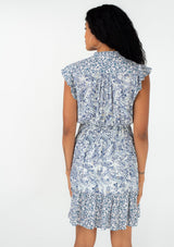 [Color: Ivory/Blue] A back facing image of a brunette model wearing a bohemian spring mini dress in a blue mixed floral and paisley print. With short ruffled cap sleeves, a ruffled neckline, a button front, a smocked elastic waist, and a tiered mini skirt. 