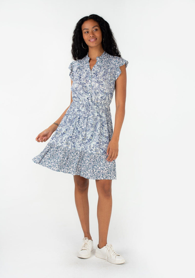 [Color: Ivory/Blue] A front facing image of a brunette model wearing a bohemian spring mini dress in a blue mixed floral and paisley print. With short ruffled cap sleeves, a ruffled neckline, a button front, a smocked elastic waist, and a tiered mini skirt. 
