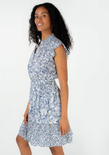 [Color: Ivory/Blue] A side facing image of a brunette model wearing a bohemian spring mini dress in a blue mixed floral and paisley print. With short ruffled cap sleeves, a ruffled neckline, a button front, a smocked elastic waist, and a tiered mini skirt. 