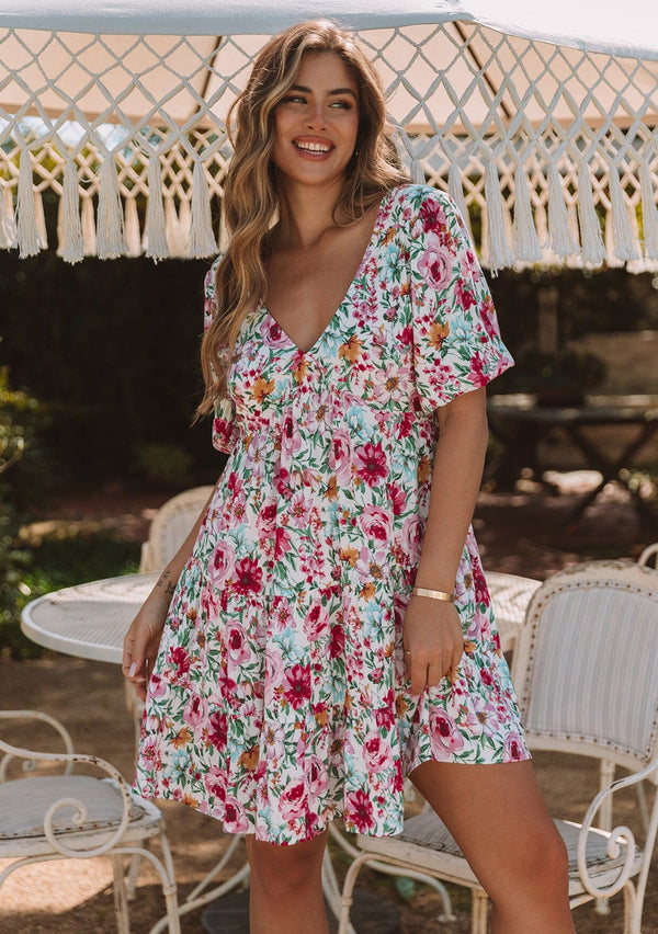[Color: Natural/Plum] A front facing image of a blonde model standing outside wearing a flowy spring mini dress in a pink floral print. With short puff sleeves, a tiered skirt, an empire waist, a v neckline in front and back, and an open back with tie closure.