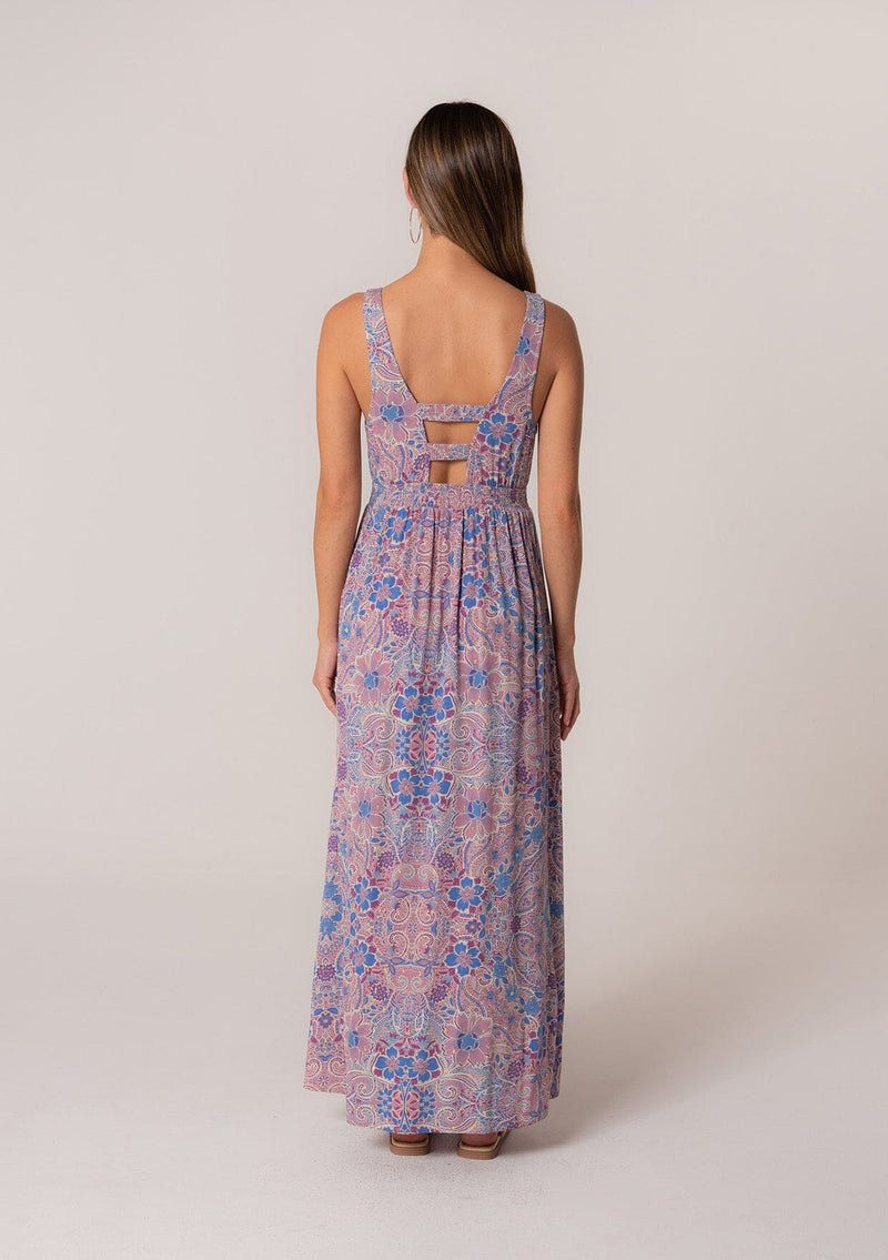 [Color: Dusty Rose/Blue] A back facing image of a brunette model wearing a bohemian summer maxi dress in a pink and blue floral print, with metallic gold clip dot details. Featuring tank top straps, a deep v neckline, a self covered button front, a front slit, a smocked elastic waist, and an open back with strap detail. 