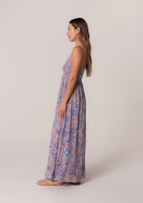 [Color: Dusty Rose/Blue] A side facing image of a brunette model wearing a bohemian summer maxi dress in a pink and blue floral print, with metallic gold clip dot details. Featuring tank top straps, a deep v neckline, a self covered button front, a front slit, a smocked elastic waist, and an open back with strap detail. 