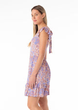 [Color: Ivory/Coral] A side facing image of a blonde model wearing a bohemian spring mini dress in a retro inspired purple floral print. With short ruffled cap sleeves, a square neckline, a ruffle trimmed tiered skirt, an open back with tie closure, and a smocked bodice at the back. 