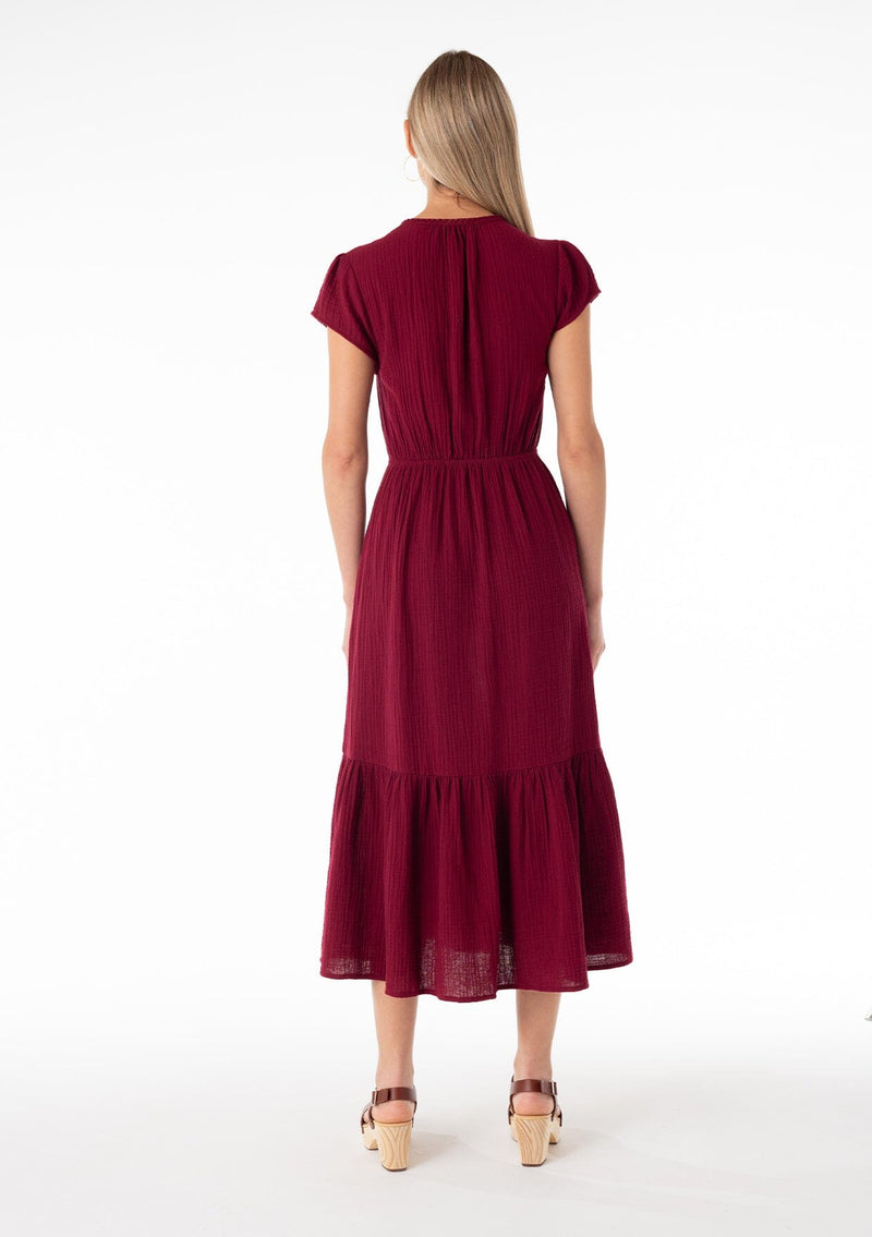 [Color: Merlot] A back facing image of a blonde model wearing a burgundy red bohemian maxi dress made with soft cotton gauze. With short cap sleeves, a tiered flowy skirt, a v neckline, a button front top, a tie waist detail, and embroidery throughout. 