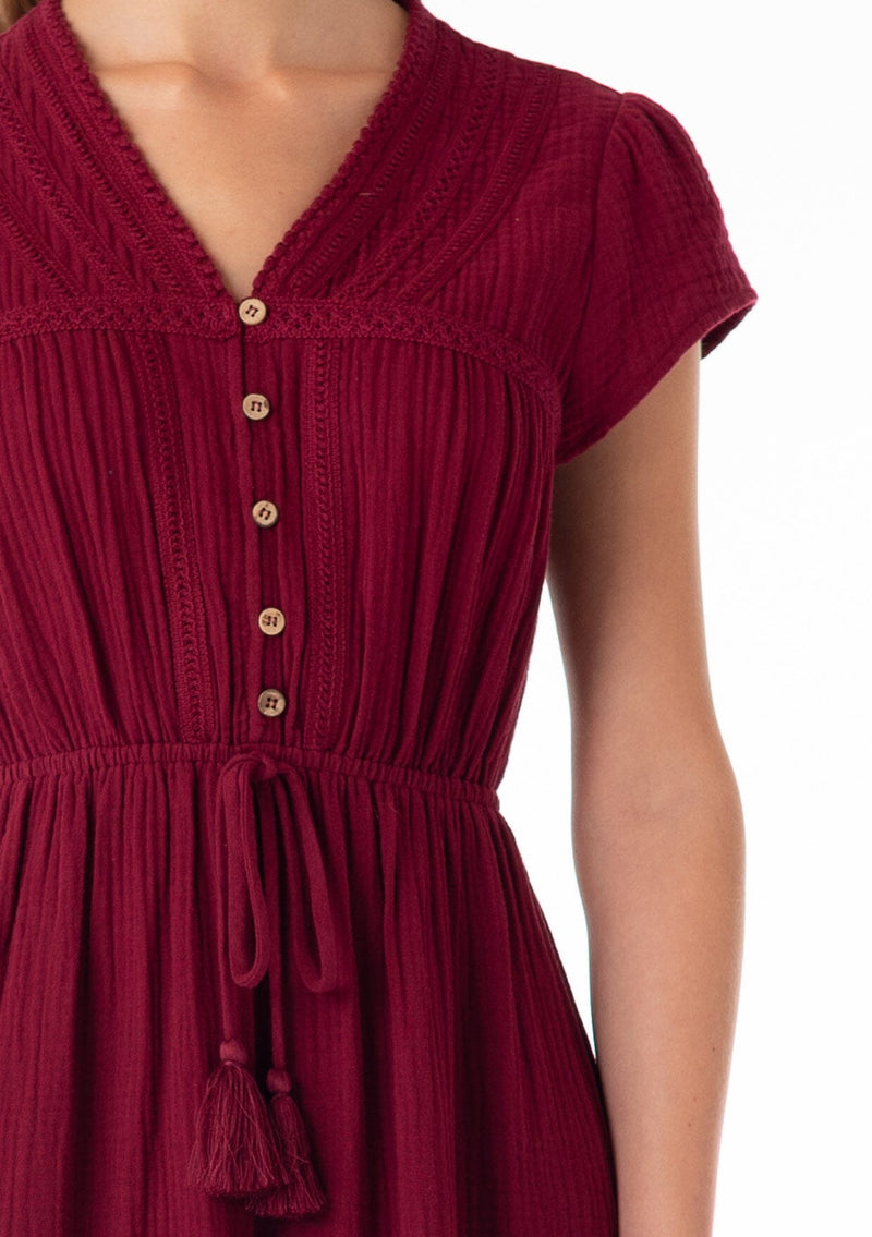 [Color: Merlot] A close up front facing image of a blonde model wearing a burgundy red bohemian maxi dress made with soft cotton gauze. With short cap sleeves, a tiered flowy skirt, a v neckline, a button front top, a tie waist detail, and embroidery throughout. 