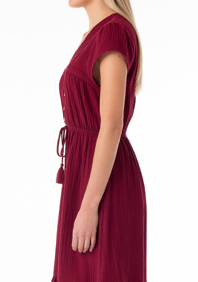[Color: Merlot] A close up side facing image of a blonde model wearing a burgundy red bohemian maxi dress made with soft cotton gauze. With short cap sleeves, a tiered flowy skirt, a v neckline, a button front top, a tie waist detail, and embroidery throughout. 