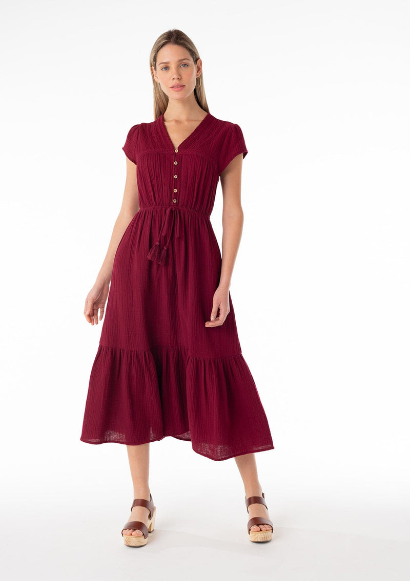 [Color: Merlot] A front facing image of a blonde model wearing a burgundy red bohemian maxi dress made with soft cotton gauze. With short cap sleeves, a tiered flowy skirt, a v neckline, a button front top, a tie waist detail, and embroidery throughout. 