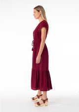 [Color: Merlot] A side facing image of a blonde model wearing a burgundy red bohemian maxi dress made with soft cotton gauze. With short cap sleeves, a tiered flowy skirt, a v neckline, a button front top, a tie waist detail, and embroidery throughout. 