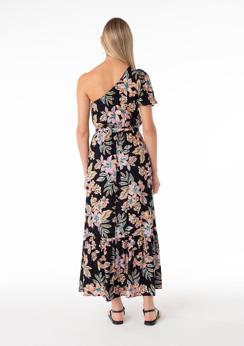 [Color: Black/Pink] A back facing image of a blonde model wearing a bohemian spring maxi dress in a tropical pink floral print. With a one shoulder top, a puff sleeve, a flowy tiered skirt, a side slit, an elastic waist, and a tie waist belt. 