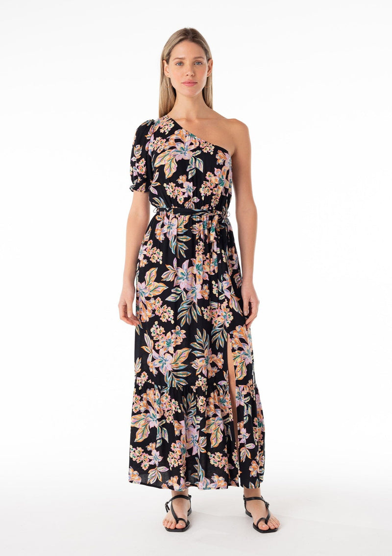 [Color: Black/Pink] A front facing image of a blonde model wearing a bohemian spring maxi dress in a tropical pink floral print. With a one shoulder top, a puff sleeve, a flowy tiered skirt, a side slit, an elastic waist, and a tie waist belt. 