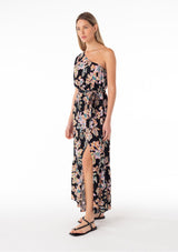 [Color: Black/Pink] A side facing image of a blonde model wearing a bohemian spring maxi dress in a tropical pink floral print. With a one shoulder top, a puff sleeve, a flowy tiered skirt, a side slit, an elastic waist, and a tie waist belt. 