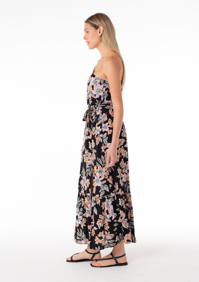 [Color: Black/Pink] A full body side facing image of a blonde model wearing a bohemian spring maxi dress in a tropical pink floral print. With a one shoulder top, a puff sleeve, a flowy tiered skirt, a side slit, an elastic waist, and a tie waist belt. 