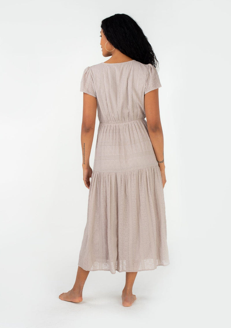 [Color: Dove] A back facing image of a brunette model wearing a light grey bohemian spring mid length dress in all over eyelet lace chiffon. With short flutter sleeves, a surplice v neckline, a tiered flowy skirt with side slit, and an elastic waist. 