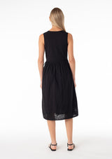 [Color: Black] A back facing image of a blonde model wearing a classic black bohemian sleeveless wrap dress designed in cotton. With a v neckline, a wrap front with side tie closure, side pockets, and embroidered detail throughout. 