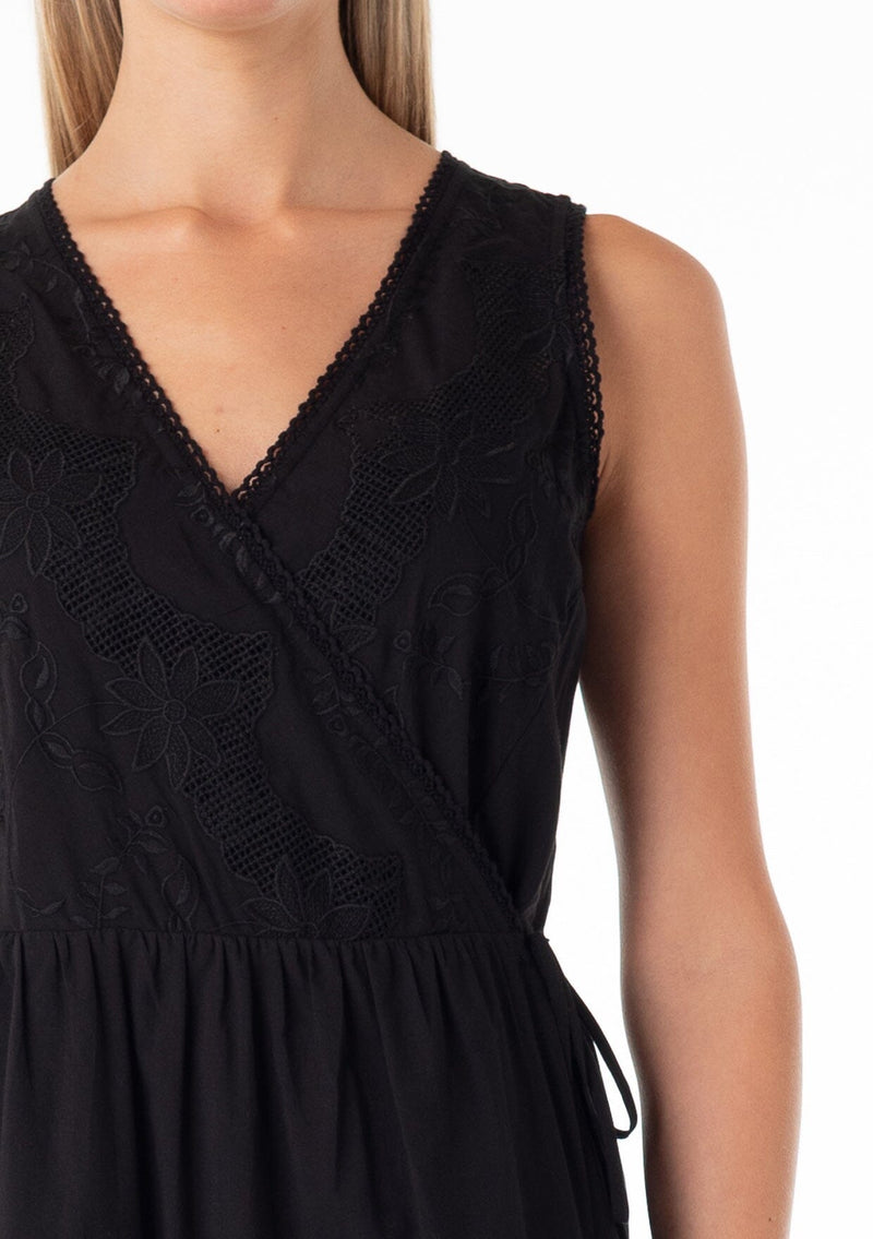 [Color: Black] A close up front facing image of a blonde model wearing a classic black bohemian sleeveless wrap dress designed in cotton. With a v neckline, a wrap front with side tie closure, side pockets, and embroidered detail throughout. 