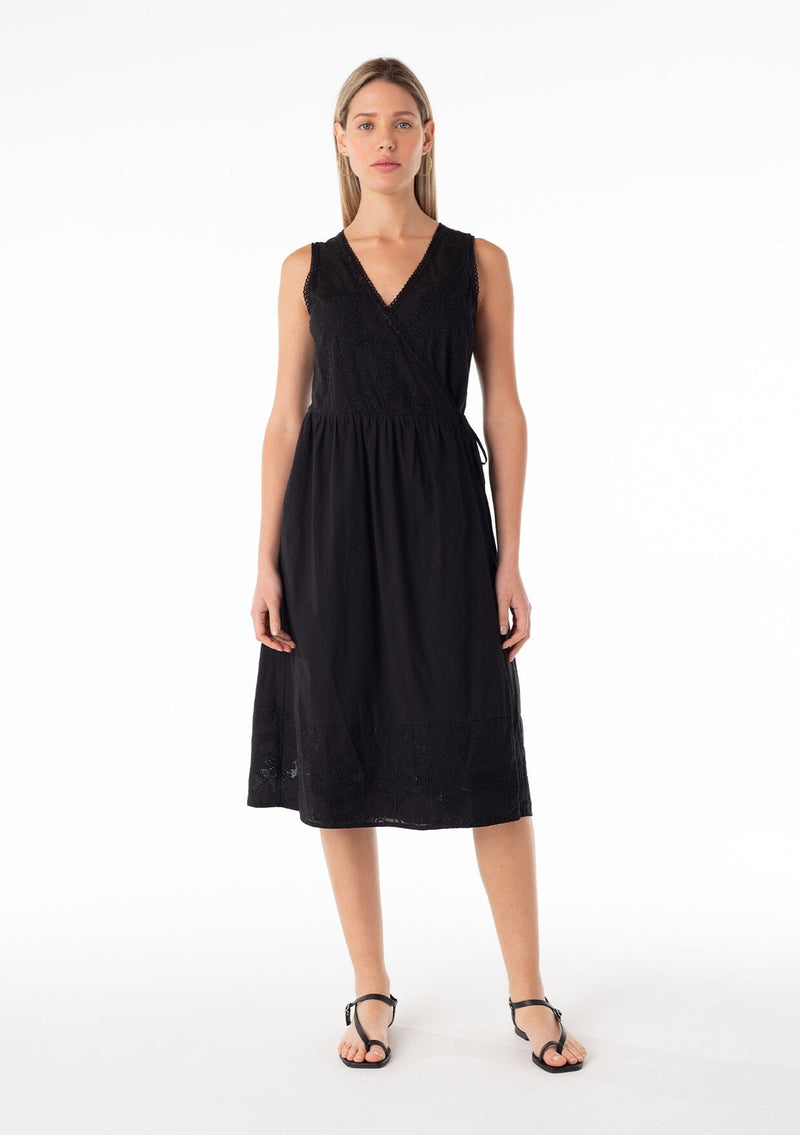 [Color: Black] A front facing image of a blonde model wearing a classic black bohemian sleeveless wrap dress designed in cotton. With a v neckline, a wrap front with side tie closure, side pockets, and embroidered detail throughout. 