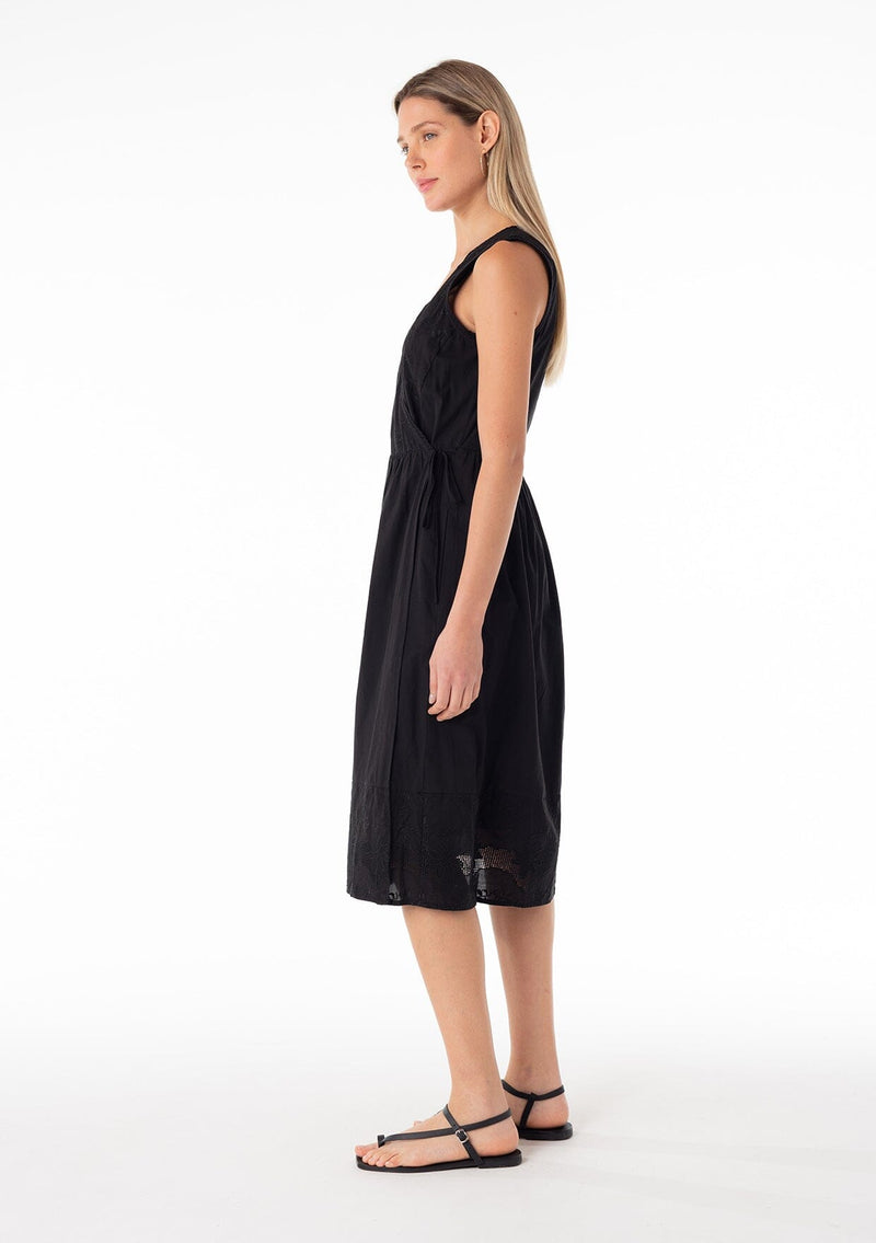 [Color: Black] A side facing image of a blonde model wearing a classic black bohemian sleeveless wrap dress designed in cotton. With a v neckline, a wrap front with side tie closure, side pockets, and embroidered detail throughout. 