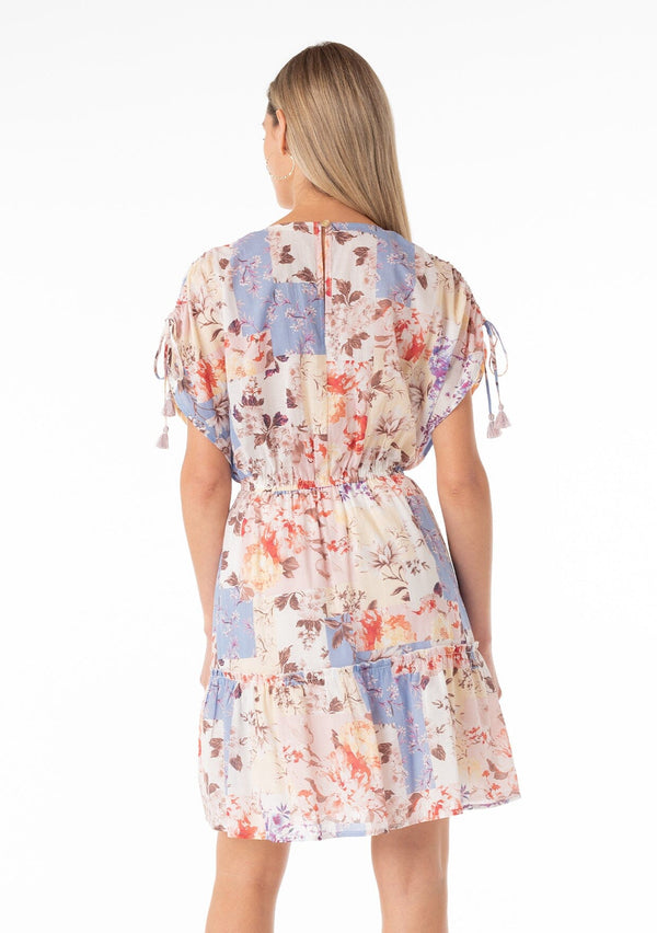 [Color: Natural/Rust] A back facing image of a blonde model wearing a bohemian spring cotton mini dress in a pink and blue floral print. With short gathered sleeves, tassel ties, a v neckline, a button front top, a ruffle trimmed tiered skirt, and a back keyhole with single button closure.
