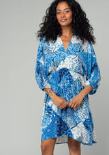 [Color: Blue/Navy] A half body front facing image of a brunette model wearing a resort bohemian mini dress in a blue patchwork floral print. With three quarter length sleeve, an open back with tassel tie closure, a high low hemline, a v neckline, and an elastic waist. 