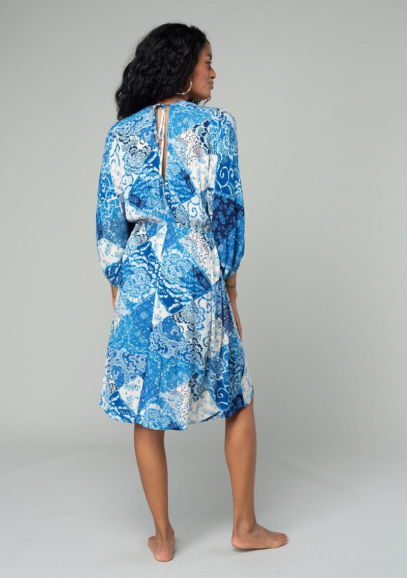 [Color: Blue/Navy] A back facing image of a brunette model wearing a resort bohemian mini dress in a blue patchwork floral print. With three quarter length sleeve, an open back with tassel tie closure, a high low hemline, a v neckline, and an elastic waist. 