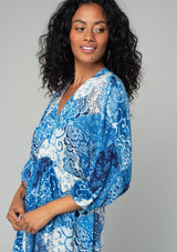 [Color: Blue/Navy] A close up side facing image of a brunette model wearing a resort bohemian mini dress in a blue patchwork floral print. With three quarter length sleeve, an open back with tassel tie closure, a high low hemline, a v neckline, and an elastic waist. 