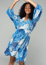 [Color: Blue/Navy] A front facing image of a brunette model wearing a resort bohemian mini dress in a blue patchwork floral print. With three quarter length sleeve, an open back with tassel tie closure, a high low hemline, a v neckline, and an elastic waist. 