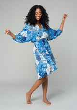 [Color: Blue/Navy] A side facing image of a brunette model wearing a resort bohemian mini dress in a blue patchwork floral print. With three quarter length sleeve, an open back with tassel tie closure, a high low hemline, a v neckline, and an elastic waist. 