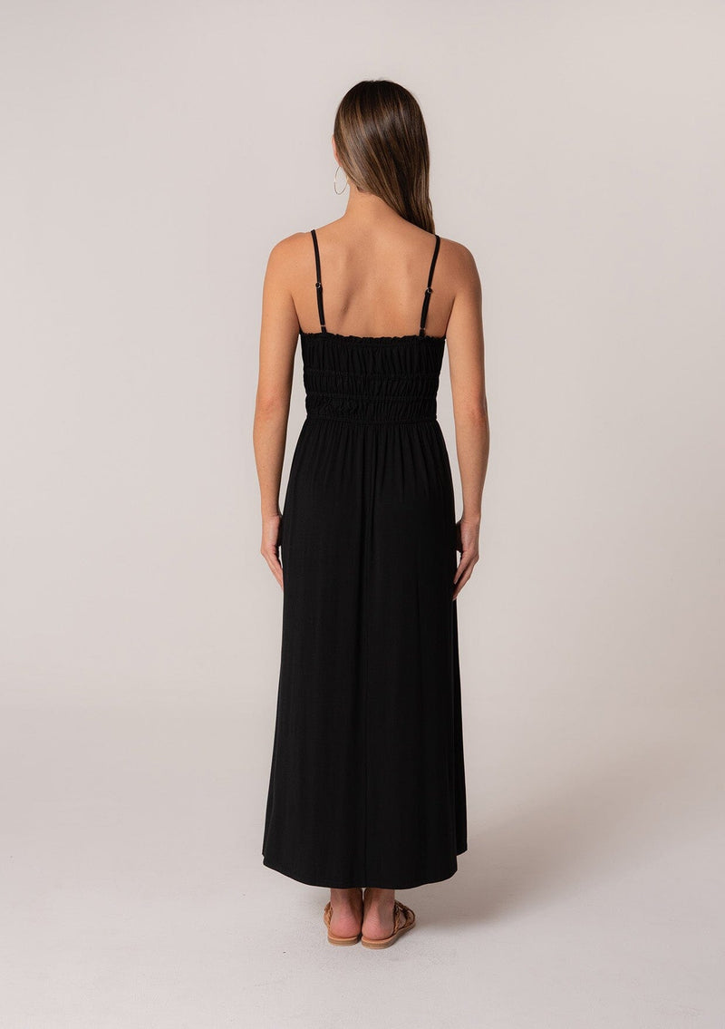 [Color: Black] A back facing image of a brunette model wearing a black sleeveless maxi dress in a stretchy bamboo knit. With spaghetti straps, a straight ruffle trimmed neckline, a smocked bodice detail, side pockets, and a long flowy skirt. 