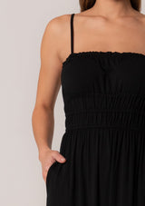 [Color: Black] A close up front facing image of a brunette model wearing a black sleeveless maxi dress in a stretchy bamboo knit. With spaghetti straps, a straight ruffle trimmed neckline, a smocked bodice detail, side pockets, and a long flowy skirt. 