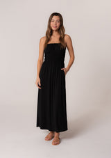 [Color: Black] A front facing image of a brunette model wearing a black sleeveless maxi dress in a stretchy bamboo knit. With spaghetti straps, a straight ruffle trimmed neckline, a smocked bodice detail, side pockets, and a long flowy skirt. 