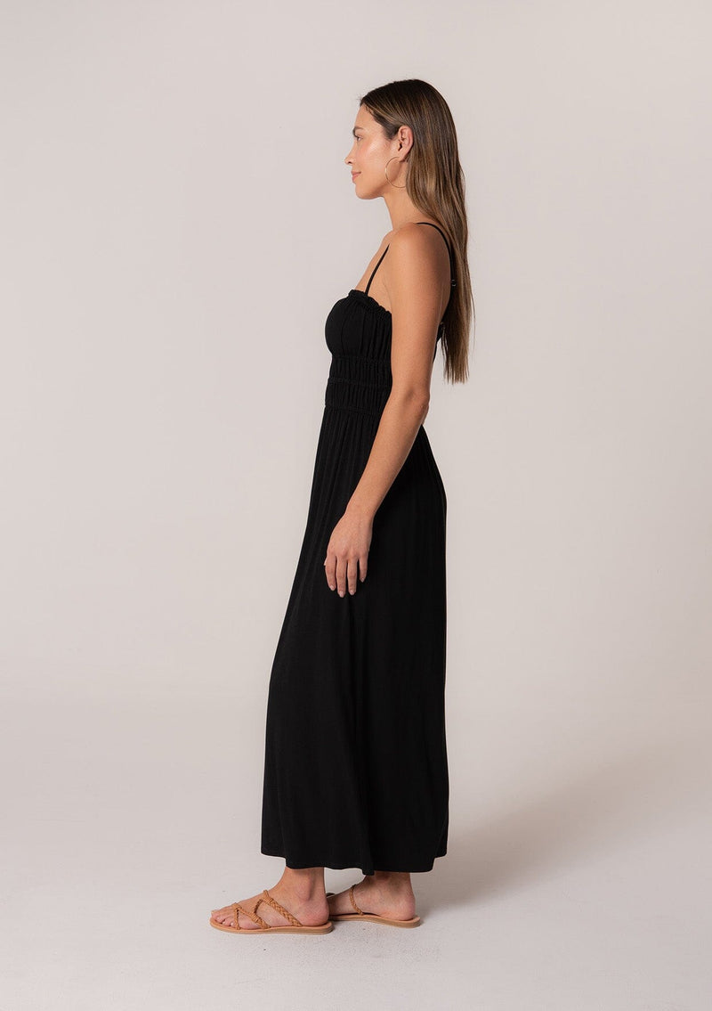 [Color: Black] A side facing image of a brunette model wearing a black sleeveless maxi dress in a stretchy bamboo knit. With spaghetti straps, a straight ruffle trimmed neckline, a smocked bodice detail, side pockets, and a long flowy skirt. 
