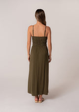 [Color: Military] A back facing image of a brunette model wearing a military green sleeveless maxi dress in a stretchy bamboo knit. With spaghetti straps, a straight ruffle trimmed neckline, a smocked bodice detail, side pockets, and a long flowy skirt. 