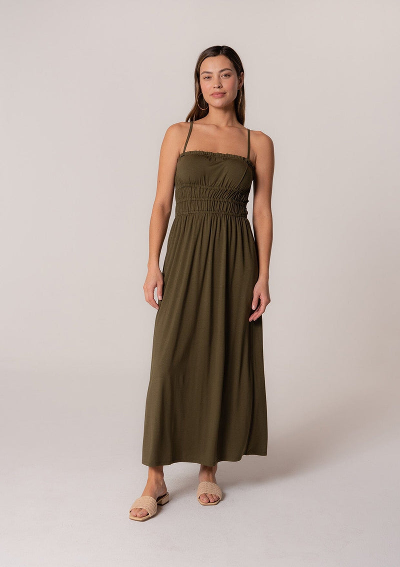 [Color: Military] A front facing image of a brunette model wearing a military green sleeveless maxi dress in a stretchy bamboo knit. With spaghetti straps, a straight ruffle trimmed neckline, a smocked bodice detail, side pockets, and a long flowy skirt. 