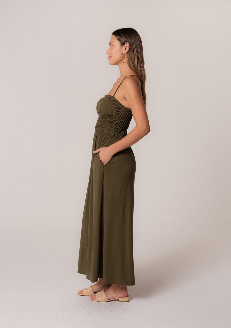 [Color: Military] A side facing image of a brunette model wearing a military green sleeveless maxi dress in a stretchy bamboo knit. With spaghetti straps, a straight ruffle trimmed neckline, a smocked bodice detail, side pockets, and a long flowy skirt. 