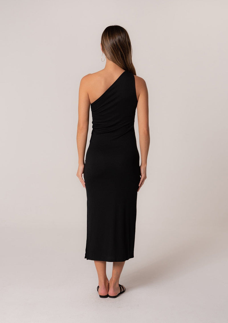 [Color: Black] A back facing image of a brunette model wearing a stretchy slim fit black knit midi dress with one shoulder strap, a cutout strap detail, and a side slit. 