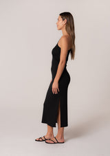 [Color: Black] A side facing image of a brunette model wearing a stretchy slim fit black knit midi dress with one shoulder strap, a cutout strap detail, and a side slit. 