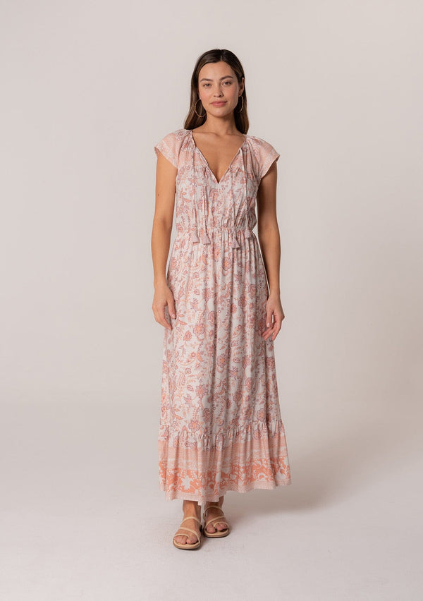 [Color: Natural/Clay] A front facing image of a brunette model wearing a pink floral bohemian maxi dress. With short cap sleeves, an elastic waist, a tiered flowy skirt, a v neckline, and double tassel tie neckline.