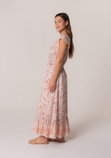 [Color: Natural/Clay] A side facing image of a brunette model wearing a pink floral bohemian maxi dress. With short cap sleeves, an elastic waist, a tiered flowy skirt, a v neckline, and double tassel tie neckline.