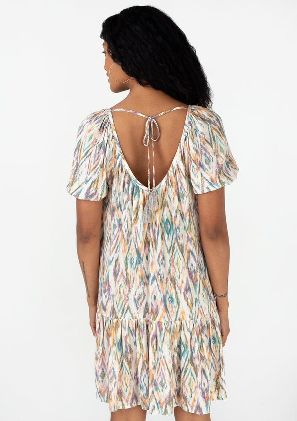 [Color: Natural/Teal] A back facing image of a brunette model wearing a bohemian resort mini dress in a multi color diamond print. With short puff sleeves, a wide v neckline, an open back with tassel tie closure, a tiered skirt, and a flowy silhouette. 