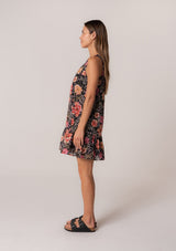 [Color: Black/Fuchsia] A side facing image of a brunette model wearing a summer bohemian cotton mini dress in a black and pink floral print. A sleeveless mini dress with a roomy silhouette, a v neckline, and a tiered skirt. 
