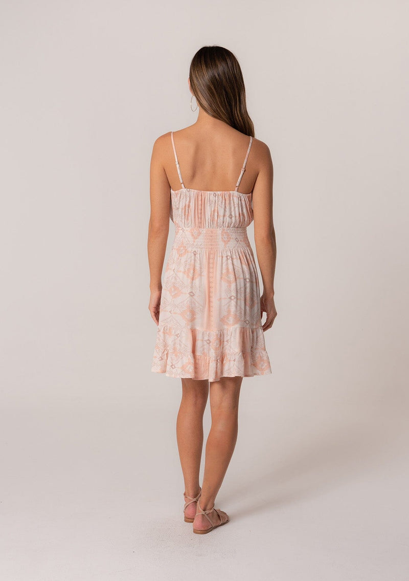 [Color: Natural/Peach] A back facing image of a brunette model wearing a summer mini dress in a pink bohemian print. With adjustable spaghetti straps, a scoop neckline, a tiered mini skirt, a smocked elastic waist, and a drawstring front detail with tassel ties. 